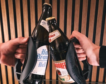 May include: Two glass beer bottles with black handles are being held up in a cheers. The bottles are both brown glass and have labels with German text. The bottles are being held in front of a wood panel wall.