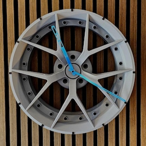 Könnte beinhalten: Eine silberfarbene Uhr mit Felgendesign und blauen Zeigern. Die Uhr hat eine kreisförmige Form mit mehreren Speichen und ist an einer Holzwand mit vertikalen Streifen befestigt.