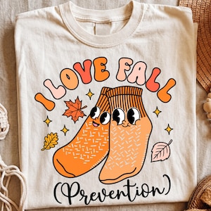May include: A cream-colored t-shirt featuring a graphic of two orange socks with cartoon faces. The text reads I LOVE FALL above the socks and (Prevention) below them. The design includes small orange and pink autumn leaves and yellow stars.