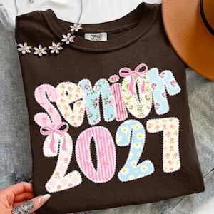 May include: A dark brown t-shirt featuring the text "SENIOR 2027" in large, decorative letters. Each letter is filled with various pastel patterns, including floral prints, stripes, and small bows. The design includes pink ribbon accents.