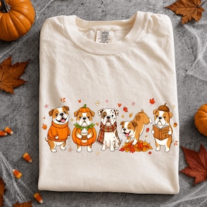 May include: A cream-colored t-shirt featuring a graphic of five cartoon bulldogs in autumn-themed attire. The dogs are depicted wearing sweaters, a pumpkin costume, a scarf, playing in leaves, and holding a book labeled Fall Reads.