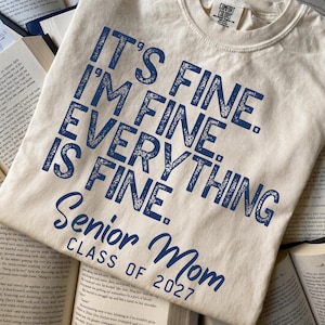 May include: A cream-colored short-sleeve t-shirt featuring dark blue distressed text that reads: IT'S FINE. I'M FINE. EVERYTHING IS FINE. Senior Mom CLASS OF 2027. The shirt has a crew neckline and a Comfort Colors brand label at the inner collar.