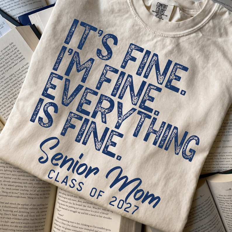 May include: A cream-colored short-sleeve t-shirt featuring dark blue distressed text that reads: IT'S FINE. I'M FINE. EVERYTHING IS FINE. Senior Mom CLASS OF 2027. The shirt has a crew neckline and a Comfort Colors brand label at the inner collar.