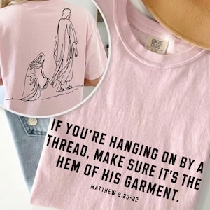 May include: A light pink t-shirt featuring a black line art illustration of a figure reaching toward another on the back. The front displays the black text: IF YOU'RE HANGING ON BY A THREAD, MAKE SURE IT'S THE HEM OF HIS GARMENT. MATTHEW 9:20-22.