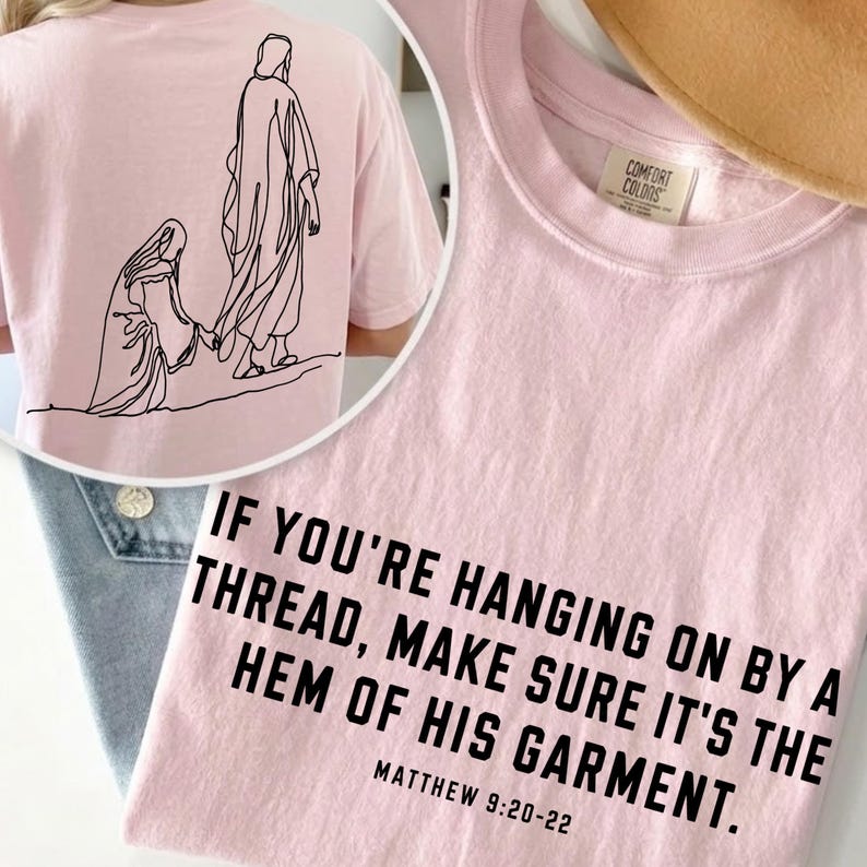 May include: A light pink t-shirt featuring a black line art illustration of a figure reaching toward another on the back. The front displays the black text: IF YOU'RE HANGING ON BY A THREAD, MAKE SURE IT'S THE HEM OF HIS GARMENT. MATTHEW 9:20-22.