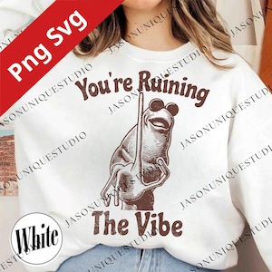 May include: White sweatshirt featuring a brown frog graphic wearing sunglasses and holding a stick. The text "You're Ruining The Vibe" is above and below the frog illustration. The sweatshirt has a crew neck.