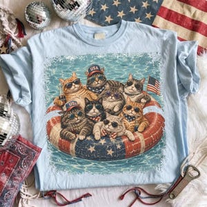 May include: Light blue t-shirt featuring a graphic of several cats wearing sunglasses and patriotic accessories, floating on a red, white, and blue inner tube. The cats are in a pool of water, with a small American flag.
