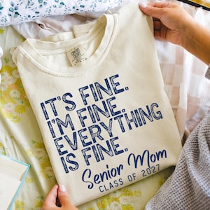 May include: A cream-colored short-sleeve t-shirt with dark blue text that reads: IT'S FINE. I'M FINE. EVERYTHING IS FINE. Senior Mom CLASS OF 2027. The shirt features a crew neckline and a Comfort Colors brand label on the inside of the collar.