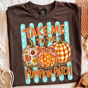 Puede incluir: Una camiseta marrón oscuro con un gráfico de tres calabazas frente a rayas verticales azul claro con lunares blancos. El texto "TAKE ME TO THE PUMPKIN PATCH" está en naranja. Las calabazas tienen patrones florales, de lunares y de cuadros.