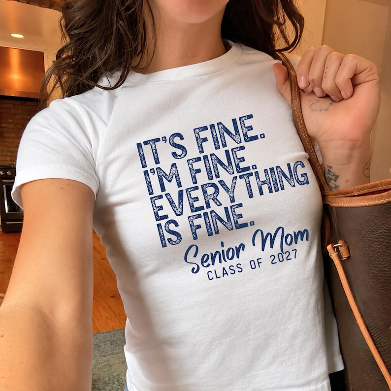May include: A white short-sleeved t-shirt featuring dark blue distressed-style text that reads: IT'S FINE. I'M FINE. EVERYTHING IS FINE. Senior Mom CLASS OF 2027. The shirt is worn with a brown leather bag strap visible over the shoulder.