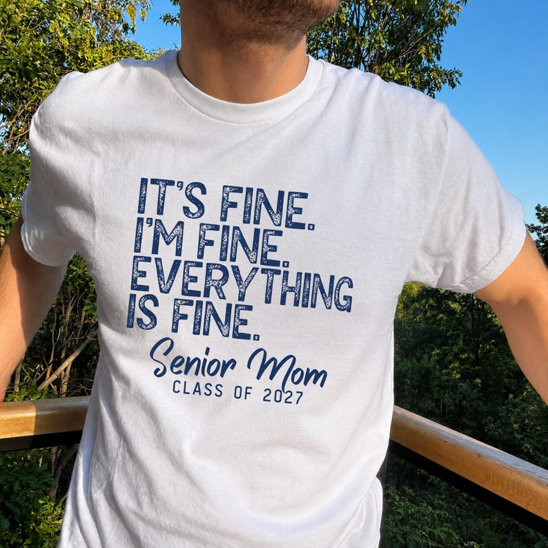 May include: A white short-sleeved t-shirt featuring dark blue text that reads: IT'S FINE. I'M FINE. EVERYTHING IS FINE. Senior Mom CLASS OF 2027. The shirt has a crew neckline and a standard fit, with the graphic printed clearly on the front chest area.
