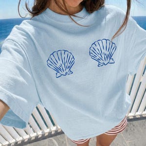 May include: A light blue short-sleeve t-shirt featuring two dark blue graphic prints of seashells positioned on the chest area. The shirt has a crew neckline and a relaxed fit, made from a solid-colored fabric with a smooth, consistent texture.