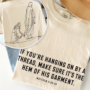 May include: A cream-colored t-shirt featuring black text that reads: IF YOU'RE HANGING ON BY A THREAD, MAKE SURE IT'S THE HEM OF HIS GARMENT. MATTHEW 9:20-22. The back of the shirt displays a black line art illustration of two figures.