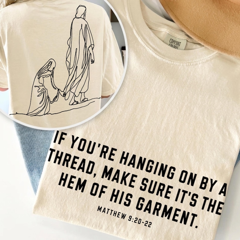 May include: A cream-colored t-shirt featuring black text that reads: IF YOU'RE HANGING ON BY A THREAD, MAKE SURE IT'S THE HEM OF HIS GARMENT. MATTHEW 9:20-22. The back of the shirt displays a black line art illustration of two figures.