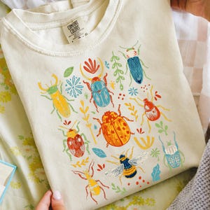 Puede incluir: Una camiseta de color crema con un estampado gráfico colorido de varios escarabajos, insectos y pequeños elementos botánicos. El diseño incluye ilustraciones de escarabajos amarillos, azules, rojos y naranjas, junto con una abeja en el frente.