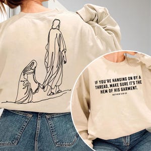 May include: A beige crewneck sweatshirt featuring a black line art illustration of a person reaching for a robe on the back, and the text "IF YOU'RE HANGING ON BY A THREAD, MAKE SURE IT'S THE HEM OF HIS GARMENT. MATTHEW 9:20-22" on the front.