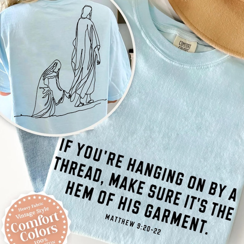May include: A light blue t-shirt featuring a black line art illustration of two figures on the back and the text "IF YOU'RE HANGING ON BY A THREAD, MAKE SURE IT'S THE HEM OF HIS GARMENT. MATTHEW 9:20-22" printed in black on the front.