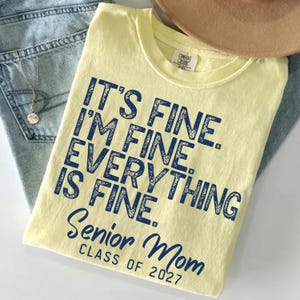 May include: A pale yellow short-sleeve t-shirt featuring dark blue distressed text that reads IT'S FINE. I'M FINE. EVERYTHING IS FINE. SENIOR MOM CLASS OF 2027. The shirt has a crew neckline and a Comfort Colors brand label on the inner collar.