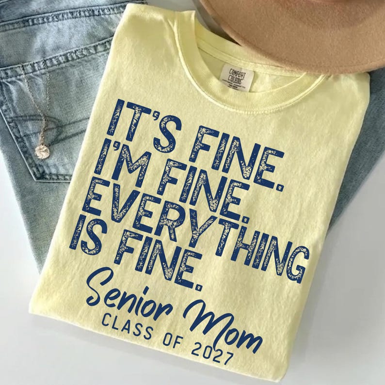 May include: A pale yellow short-sleeve t-shirt featuring dark blue distressed text that reads IT'S FINE. I'M FINE. EVERYTHING IS FINE. SENIOR MOM CLASS OF 2027. The shirt has a crew neckline and a Comfort Colors brand label on the inner collar.