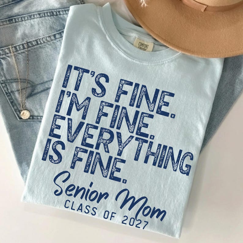 May include: A light blue t-shirt featuring dark blue text that reads: IT'S FINE. I'M FINE. EVERYTHING IS FINE. Senior Mom CLASS OF 2027. The shirt has a crew neckline and a tag near the collar. The text has a distressed, textured appearance.
