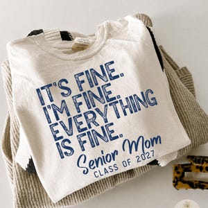 May include: A cream-colored t-shirt featuring dark blue distressed text that reads IT'S FINE. I'M FINE. EVERYTHING IS FINE. SENIOR MOM CLASS OF 2027. The shirt is layered over a beige and black textured knit garment, with a tortoiseshell hair clip nearby.