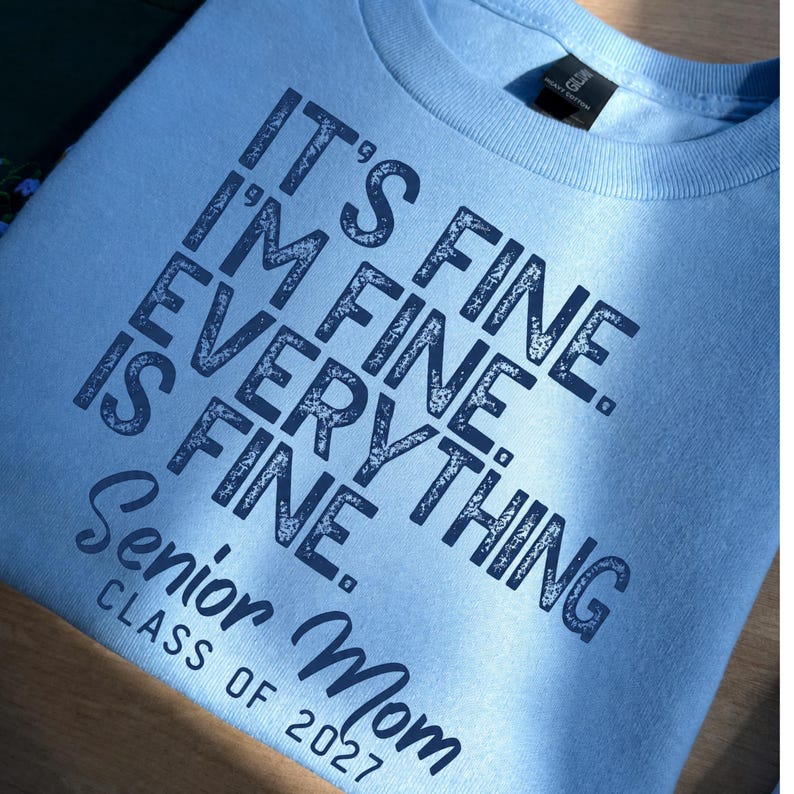 May include: A light blue t-shirt featuring dark navy blue text that reads: IT'S FINE. I'M FINE. EVERYTHING IS FINE. SENIOR MOM CLASS OF 2027. The lettering has a distressed, textured appearance, and the shirt includes a black neck label.