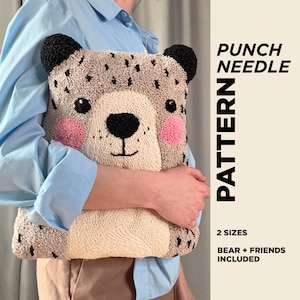 Op de afbeelding: Een handgemaakt, getextureerd kussen in de vorm van een beer met zwarte oren, zwarte stippen en roze wangen. Het kussen heeft een crèmekleurig gezicht. De tekst rechts luidt "PUNCH NEEDLE PATTERN" en "2 MATEN BEAR + FRIENDS INCLUDED." Een persoon draagt een lichtblauw overhemd.