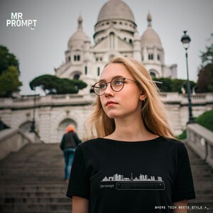 Op de afbeelding: Een zwart t-shirt met een witte skyline-afbeelding en de tekst "/prompt" wordt gedragen door een persoon met blond haar en een bril. De zin "WHERE TECH MEETS STYLE" staat onderaan. Op de achtergrond is een kerk te zien.