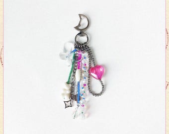 May include: A silver keychain with a crescent moon charm, adorned with a variety of charms. These include a pink heart, clear and colorful beads, a white flower, and a small white bear. The keychain has a decorative chain.