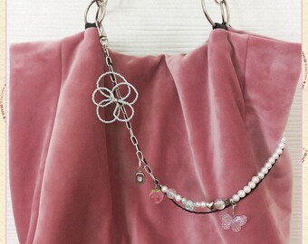 May include: A dusty rose velvet tote bag with silver metal handles. A decorative chain with a white beaded flower, a pink strawberry charm, and a clear butterfly charm hangs from the handle.