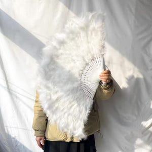 May include: A large, white feather fan. The fan is held open, obscuring the person holding it. The feathers are fluffy and soft, and the fan's handle is white. The background is a white curtain.