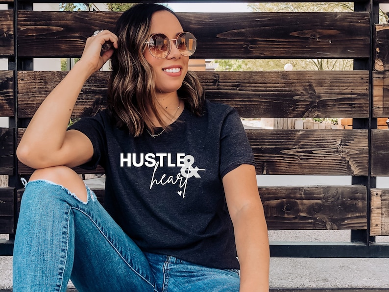 May include: A woman wearing a black t-shirt with the text "Hustle & heart" in white lettering. She is sitting on a wooden bench and wearing blue jeans and sunglasses.