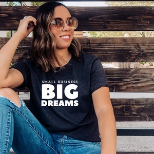 May include: A woman wearing a dark grey t-shirt with the text "SMALL BUSINESS BIG DREAMS" in white. She is sitting on a wooden bench and wearing blue jeans.