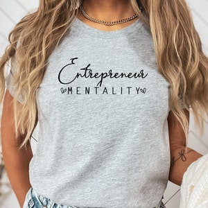 May include: A grey t-shirt with the text "Entrepreneur Mentality" in black script lettering with a heart on either side of the word "Mentality".