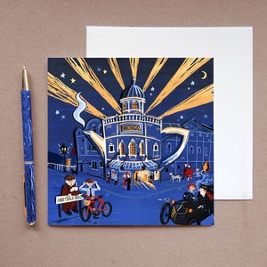 Puede incluir: Una tarjeta de felicitación caprichosa con un cine diseñado como una tetera, con una combinación de colores azul y dorado. La tarjeta incluye un bolígrafo azul a juego con un patrón de ondas. La escena representa una calle con gente, una bicicleta y un coche antiguo.