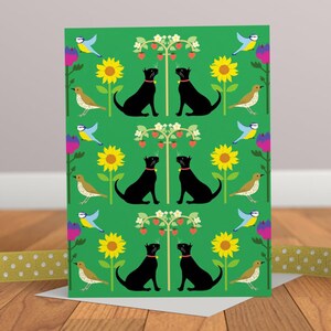May include: Greeting card with a green background featuring a repeating pattern of black cats, sunflowers, birds, and strawberry plants. The cats wear red collars. The card is on a wooden surface with a yellow ribbon.