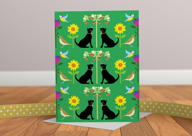 May include: Greeting card with a green background featuring a repeating pattern of black cats, sunflowers, birds, and strawberry plants. The cats wear red collars. The card is on a wooden surface with a yellow ribbon.
