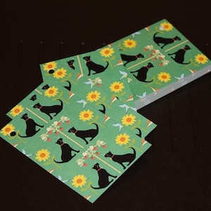 May include: A stack of green square paper coasters with a repeating pattern of black cats wearing red collars, yellow sunflowers, and blue birds. The coasters are stacked on a dark surface.