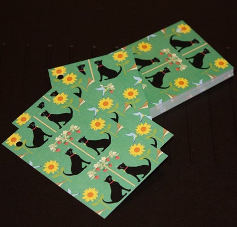 May include: A stack of green square paper coasters with a repeating pattern of black cats wearing red collars, yellow sunflowers, and blue birds. The coasters are stacked on a dark surface.
