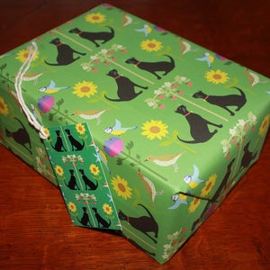May include: A wrapped gift box with green wrapping paper featuring a pattern of black dogs, sunflowers, birds, and floral elements. A matching gift tag is attached with twine. The overall theme is a nature-inspired gift.