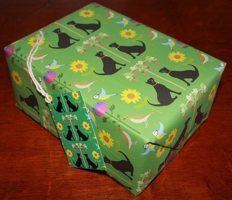 May include: A wrapped gift box with green wrapping paper featuring a pattern of black dogs, sunflowers, birds, and floral elements. A matching gift tag is attached with twine. The overall theme is a nature-inspired gift.