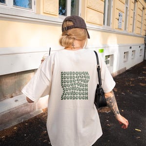 May include: A person wearing a white t-shirt with the word "SOURDOUGH" repeated in green text on the back. The person is wearing a black baseball cap and a black bag.