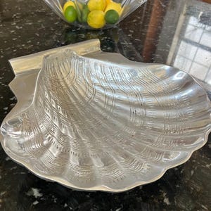 Könnte beinhalten: Eine silberfarbene Metallschale in Form einer großen Jakobsmuschel. Die Oberfläche weist ein strukturiertes, geripptes Design mit komplexen, sich wiederholenden Mustern entlang der geschwungenen Kanten auf, was eine reflektierende Oberfläche erzeugt.