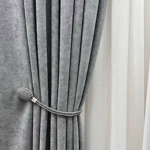 Puede incluir: Una cortina gris con un amarre de cuerda plateado. El amarre tiene una bola decorativa en un extremo.