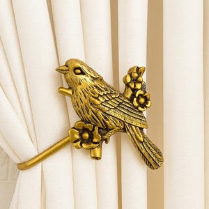 May include: A golden-colored metal curtain tieback shaped like a bird perched on a branch with flowers. The bird is detailed with textured feathers and is holding the curtain in place.