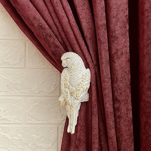 May include: A decorative parrot curtain tieback in a cream and gold finish. The parrot is attached to a deep red velvet curtain. The tieback is mounted on a white brick wall. Home decor.