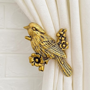 May include: A gold-colored metal curtain tieback in the shape of a bird perched on a flowering branch. The bird is detailed with textured feathers and is holding a branch with flowers. The tieback is holding a white curtain.