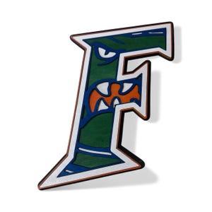 May include: A wooden letter "F" in a stylized design, featuring a green background with a blue and orange monster face. The letter has a white border and a brown outline, creating a 3D effect. The design is likely for a sports team or school.