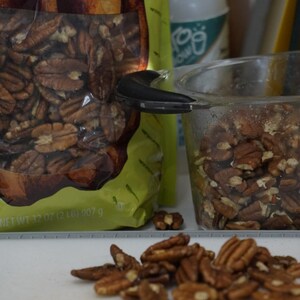 May include: A bag of pecan halves with the text "Pecans" on the front. The bag is open and some pecans are spilling out. There is a clear glass measuring cup filled with pecans in the background.