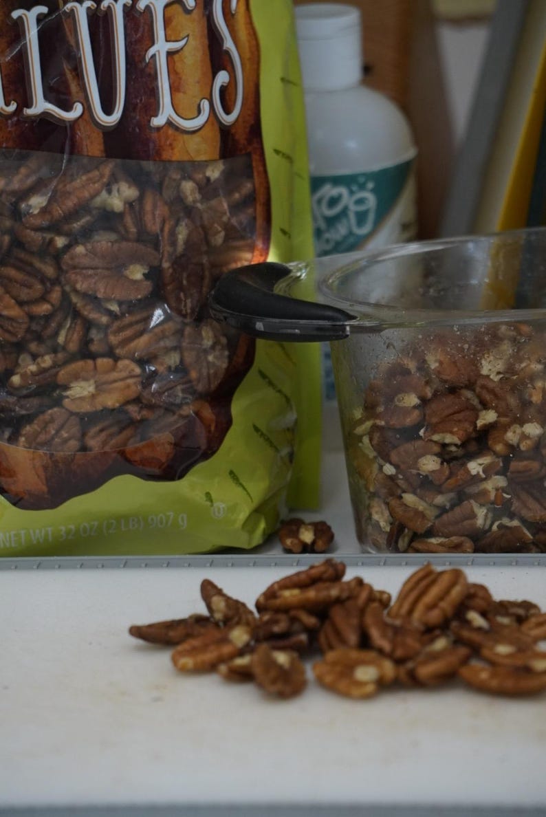 May include: A bag of pecan halves with the text "Pecans" on the front. The bag is open and some pecans are spilling out. There is a clear glass measuring cup filled with pecans in the background.